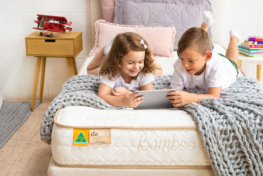 Two children lying on a bamboo mattress, using a tablet in a bright bedroom setting